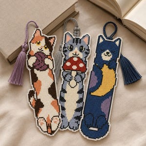 Puede incluir: Tres marcapáginas verticales de punto de cruz con diseños de gatos. El primero es un gato calicó que sostiene un ovillo de lana morado con una borla lavanda. El segundo es un gato atigrado gris con una seta roja y una borla gris. El tercero es un gato azul oscuro con una luna creciente amarilla y una borla azul oscuro.