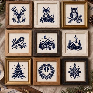 May include: A collection of nine square framed cross-stitch artworks featuring dark blue thread designs on cream fabric. The motifs include a deer, fox, owl, bird, mountain landscape, cabin, Christmas tree, wreath, and snowflake in various wood frames.
