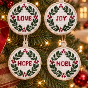 May include: Four circular cross-stitch ornaments hanging on a tree. Each ornament features a white fabric center with a green wreath and red berries. The words LOVE, JOY, HOPE, and NOEL are stitched in red in the center of each respective wreath.