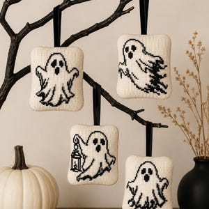 Puede incluir: Cuatro adornos rectangulares de tela color crema con diseños de fantasmas en punto de cruz negro. Cada adorno cuelga de una cinta negra. Los fantasmas aparecen en varias posturas, incluyendo uno que sostiene un farol, con detalles bordados en negro.