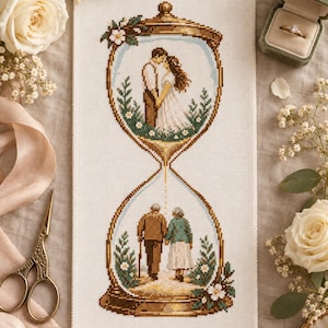 May include: A cross-stitch embroidery on white fabric depicting an hourglass. The top bulb shows a young couple standing together, while the bottom bulb shows an elderly couple walking away. The frame is brown with green foliage and white flowers.