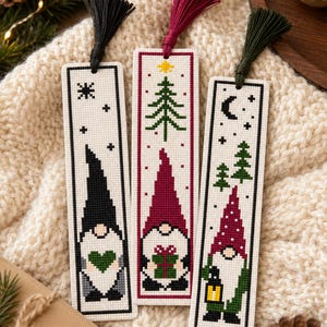 May include: Three rectangular cross-stitch bookmarks on a cream knit fabric. Each features a gnome: one with a black hat and green heart, one with a maroon hat and gift, and one with a maroon hat, lantern, and pine trees. Each has a colored tassel.