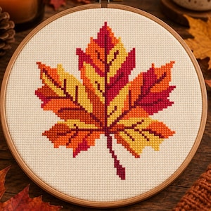 Peut inclure: Un projet de broderie au point de croix représentant une grande feuille d'érable dans des tons de rouge, orange et jaune, centré sur un tissu Aida blanc cassé. Le tissu est tendu dans un tambour à broder circulaire en bois avec une vis.