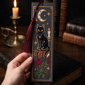 Puede incluir: Un marcapáginas rectangular con un diseño de punto de cruz sobre un fondo gris oscuro. El patrón muestra una luna creciente, un gato negro sentado sobre un libro junto a una vela encendida y una rosa roja. Una borla roja oscura cuelga arriba.