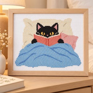 Könnte beinhalten: Eine Kreuzstichstickerei in einem hellen Holzrahmen, die eine schwarze Katze mit gelben Augen zeigt, die im Bett ein rotes Buch liest. Die Katze ist mit einer blauen Decke bedeckt und lehnt an einem cremefarbenen Kissen mit einem kleinen rosa Kissen.