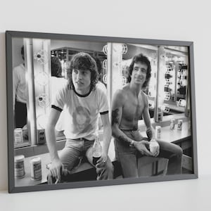 May include: A black and white framed photograph depicting two individuals in a dressing room with vanity lights. One individual wears a ringer t-shirt and jeans, holding a glass bottle. The other is shirtless with arm tattoos, holding a cup.