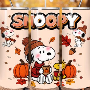 May include: A cylindrical tumbler with a light peach background featuring Snoopy and Woodstock characters, autumn leaves, pumpkins, and acorns. The word SNOOPY is printed in large, bold, orange-outlined letters at the top. Includes a metal straw.