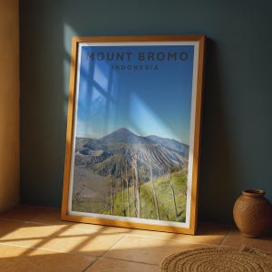 Puede incluir: Una impresión de arte mural vertical enmarcada que presenta una fotografía de paisaje del Monte Bromo, Indonesia. La imagen muestra una cadena montañosa volcánica bajo un cielo azul claro, con árboles secos sin hojas y el texto MOUNT BROMO INDONESIA.