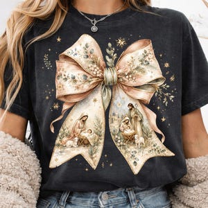 Könnte beinhalten: Ein anthrazitfarbenes T-Shirt mit einer großen, zentralen Grafik einer beigen Schleife. Die Schleife ist mit botanischen Elementen und zwei Krippenszenen verziert, die Figuren, ein Baby in einer Krippe und Tiere zeigen, umgeben von kleinen Goldsternen.