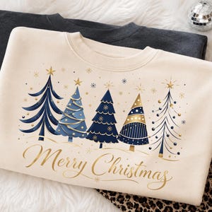 May include: A cream-colored crewneck sweatshirt featuring a graphic of five stylized Christmas trees in shades of navy blue, light blue, and gold. The text Merry Christmas is printed in gold cursive script below the trees, accented by gold snowflakes.