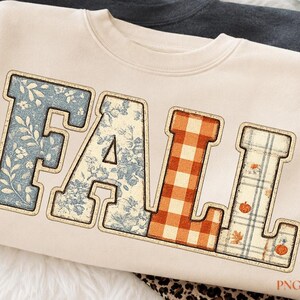Peut inclure: Un sweat-shirt à col rond couleur crème portant le mot FALL en grandes lettres majuscules. Chaque lettre présente un motif unique : le F et le A ont un imprimé floral bleu, le L a un motif vichy orange et le L un motif écossais avec des citrouilles.