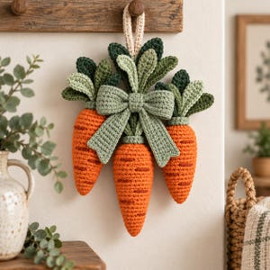 Pode incluir: Um adorno de parede com três cenouras de crochê laranja com folhas verdes texturizadas, amarradas com um grande laço de crochê verde-sálvia. O conjunto está suspenso por uma pequena alça de crochê na cor creme.