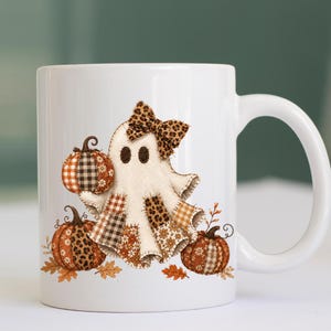 Może przedstawiać: Biały ceramiczny kubek z grafiką ducha noszącego kokardę w panterkę. Duch jest ozdobiony łatami w kratę, kwiaty i wzory panterki. Trzy dynie z podobnymi wzorami znajdują się wokół ducha na powierzchni białego kubka.