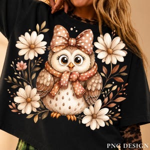 May include: A black sweatshirt featuring a central graphic of a brown owl with large eyes, wearing a polka-dot bow and scarf. The owl is surrounded by white daisy flowers and brown foliage. The text PNG DESIGN is printed in the bottom right corner.