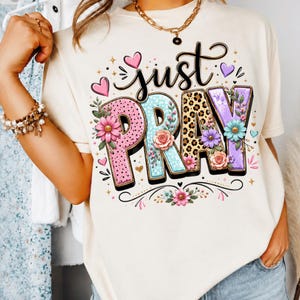 Może przedstawiać: Kremowy t-shirt z napisem "just PRAY" stylizowaną czcionką. Litery ozdobione są różowymi kropkami, jasnoniebieskimi kropkami, wzorem w panterkę i fioletowymi motywami, podkreślone kolorowymi ilustracjami kwiatów oraz małymi serduszkami.