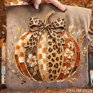 May include: A brown t-shirt featuring a textured, painted pumpkin illustration. The pumpkin has alternating sections of leopard print and a checkered pattern, adorned with a large leopard print bow and white floral accents on the sides.