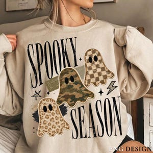 以下が含まれることがあります： ベージュのクルーネックトレーナーで、黒い文字で「SPOOKY SEASON」と書かれています。デザインには、茶色とタンの市松模様、緑の迷彩柄、茶色のヒョウ柄という、異なるパターンの3つの幽霊のイラストが描かれています。