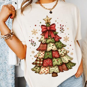 Puede incluir: Una camiseta de manga corta color crema con un gráfico grande de un árbol de Navidad hecho de patrones de tela de patchwork en rojo, verde y blanco. El árbol está rematado con una estrella dorada y un gran lazo rojo, rodeado de copos y bayas.