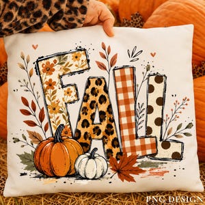 May include: A white square throw pillow featuring the word FALL in large letters. The F has a floral pattern, the A has a leopard print, the L has an orange gingham check, and the L has brown polka dots. Two pumpkins and leaves surround the text.