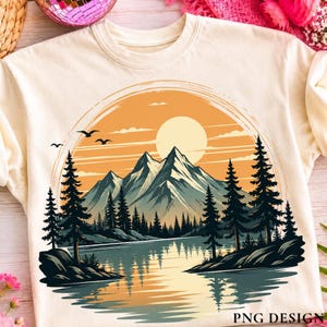 May include: A cream-coloured t-shirt featuring a circular graphic of a mountain range, a lake, and a forest of pine trees under a large orange sun. The design includes silhouettes of birds flying in the sky and the text PNG DESIGN at the bottom.