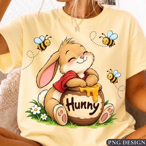 May include: A pale yellow t-shirt featuring a graphic of a brown rabbit wearing a red shirt, leaning on a brown honey pot labelled "Hunny" with dripping honey. Three small bees fly around the rabbit, and white flowers grow at the base.