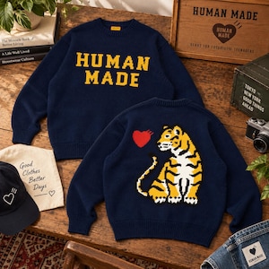 Puede incluir: Un suéter de punto azul marino de manga larga con el texto HUMAN MADE en amarillo en la parte delantera y un gráfico de un tigre con un corazón rojo en la parte posterior. El suéter tiene cuello redondo y puños y dobladillo acanalados.