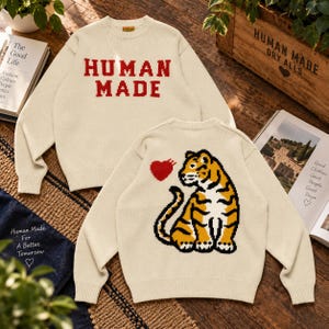 May include: A cream-coloured long-sleeve knit jumper featuring the red text HUMAN MADE on the front. The back of the jumper displays a large graphic of a seated tiger in orange, black, and white, accompanied by a small red heart icon above it.