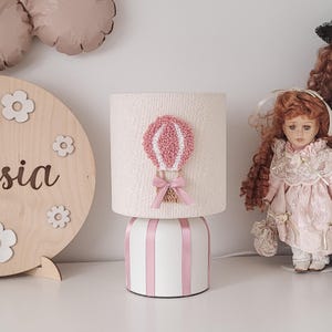 Könnte beinhalten: Eine Tischlampe mit einem zylindrischen, cremeweißen Lampenschirm, der eine texturierte Applikation eines rosa-weißen Heißluftballons mit einer rosa Schleife zeigt. Der Lampenfuß ist weiß und mit vertikalen rosa Streifen versehen.