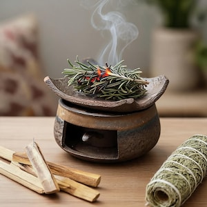 May include: A dark brown ceramic incense burner with a wide, curved top dish holding burning rosemary sprigs that emit white smoke. Beside the burner are several light-colored wooden sticks and a cylindrical bundle of dried green herbs tied with string.