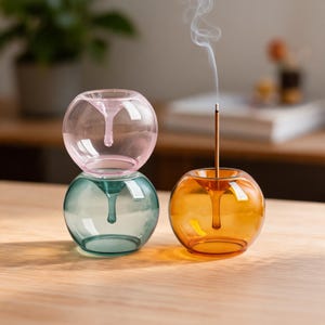 May include: Three spherical glass incense holders with a central funnel-shaped opening. One holder is light pink, one is teal, and one is amber. The amber holder contains a single upright incense stick emitting a thin trail of white smoke.