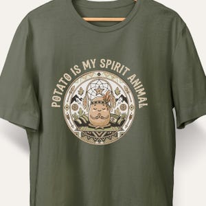 May include: An olive green t-shirt featuring a circular graphic with the text "POTATO IS MY SPIRIT ANIMAL." The design depicts a cartoon potato meditating in a lotus position, surrounded by a dreamcatcher, mountains, cacti, and geometric patterns.