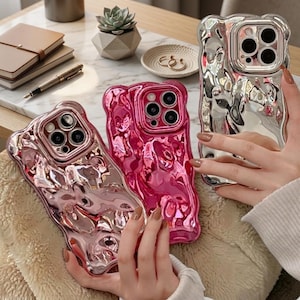 Puede incluir: Tres fundas para smartphone con un acabado metálico reflectante similar al líquido. Una funda es plateada, otra es rosa brillante y otra es de un tono oro rosa más claro. Cada funda presenta una textura irregular fundida y un recorte cuadrado.