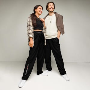 Puede incluir: Pantalones de chándal negros con una cintura elástica blanca con el texto STAFF FIGHTERS y el mismo texto impreso verticalmente en blanco en la pierna derecha. Los pantalones tienen un corte relajado y se llevan con zapatillas blancas.