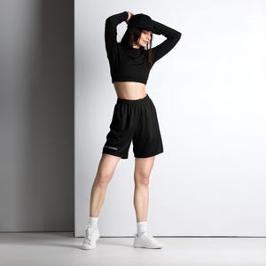 Op de afbeelding: Een zwart sportshirt met lange mouwen en blote buik, een zwarte baseballpet en een zwarte sportshort met de tekst "FOO FIGHTERS" in het wit op de rechterpijp. De outfit is gecombineerd met witte sokken en witte sportschoenen.