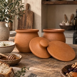 Op de afbeelding: Twee terracotta potten met bijpassende ronde deksels, voorzien van een natuurlijke matte afwerking en een ronde lichaamsvorm. De potten staan naast elkaar, waarbij één deksel rechtop staat en het andere deksel plat op het oppervlak ligt.