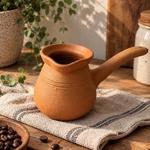 Könnte beinhalten: Eine Kaffeekanne aus Terrakotta mit bauchigem Körper, breitem Ausguss und einem langen, abgewinkelten Griff. Die Außenfläche hat ein mattes, erdbraunes Finish mit zwei dezenten horizontalen Rillen im oberen Halsbereich.