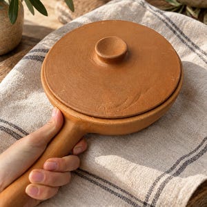 Puede incluir: Una sartén redonda de arcilla color terracota con una tapa circular a juego y un mango largo y resistente. La superficie del utensilio tiene una textura mate y terrosa, mostrando un acabado natural sin esmaltar de alfarería.