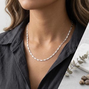 Peut inclure: Un collier chaîne en argent composé d'une série de petits maillons plats de forme ovale avec une finition texturée et réfléchissante. Le collier repose autour du cou, présentant un motif cohérent de segments métalliques interconnectés.