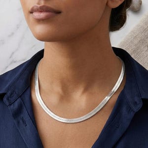 Puede incluir: Un collar de cadena tipo espiga en tono plateado con un diseño plano y flexible de acabado pulido y reflectante. El collar descansa sobre el cuello, mostrando un patrón continuo de eslabones metálicos pequeños e inclinados entrelazados.