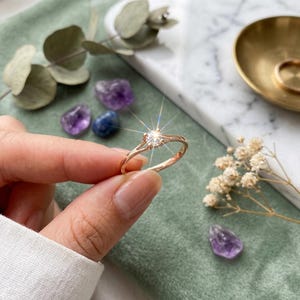May include: A rose gold ring featuring a single round brilliant-cut clear stone set in a prong setting. The band is thin and polished, reflecting light with a starburst effect at the centre of the stone, showcasing a classic solitaire design.