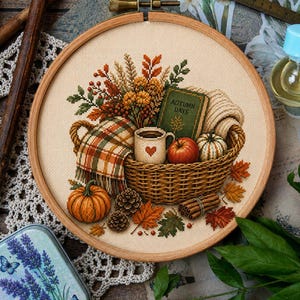 Puede incluir: Un bastidor de bordado circular de madera que contiene un diseño de punto de cruz de una cesta tejida llena de una manta a cuadros, una taza con un corazón, una manzana, un libro titulado AUTUMN DAYS, una calabaza, piñas, canela y hojas.