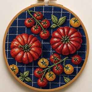 May include: A circular wooden embroidery hoop containing a cross-stitch pattern of red and yellow tomatoes on a blue and white chequered fabric background. The design features two large tomatoes and several smaller clusters of tomatoes and green leaves.