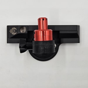 May include: A black metal mounting bracket featuring a central red cylindrical component with small circular vents at the top. The bracket includes a black adjustment knob and a small engraved logo on the left side of the horizontal base plate.