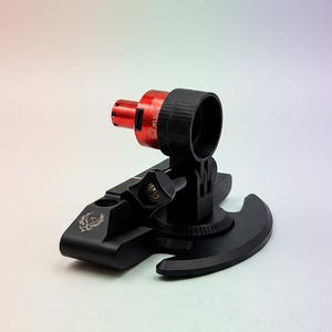 Może przedstawiać: Czarno-czerwone mechaniczne urządzenie montażowe z okrągłą podstawą, regulowanym ramieniem z pokrętłem zaciskowym oraz cylindrycznym czerwonym elementem z małym portem. Na podstawie znajduje się mały wygrawerowany emblemat ptaka z rozpostartymi skrzydłami.