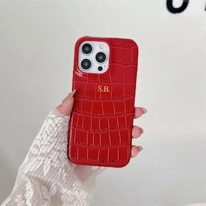 Puede incluir: Una funda de smartphone de color rojo brillante con un patrón texturizado de piel de cocodrilo. La funda cuenta con un recorte para un sistema de cámara de triple lente y las iniciales S.B. doradas grabadas en el centro.