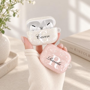 May include: Two protective cases for wireless earbuds featuring a pearlescent finish. One case is white with the name Caren and a large letter C, while the other is light pink with the name Randa and a large letter R printed in black script.