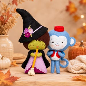 May include: Two felt plush dolls: one is a green-faced witch wearing a black pointed hat with purple flowers and a lavender dress, holding a yellow broom; the other is a blue monkey with a red hat and vest, standing next to the witch.