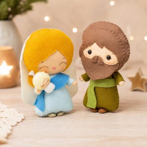 May include: Two handmade felt dolls representing a nativity scene. One doll has yellow hair, a white head covering, and a light blue dress, holding a small white-wrapped infant. The other doll has a brown beard, brown hair, and a dark olive green tunic.