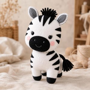 May include: A handcrafted felt zebra plush toy featuring a white body with black stripes, a black muzzle, and a black yarn mane and tail. The zebra has stitched black eyes, pink circular cheeks, and grey inner ears with visible white stitching.