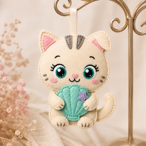 May include: A cream-colored felt cat ornament featuring large teal eyes, pink cheeks, and a small pink nose. The cat holds a teal seashell with a small purple star attached to the center. It has a cream ribbon loop at the top for hanging.